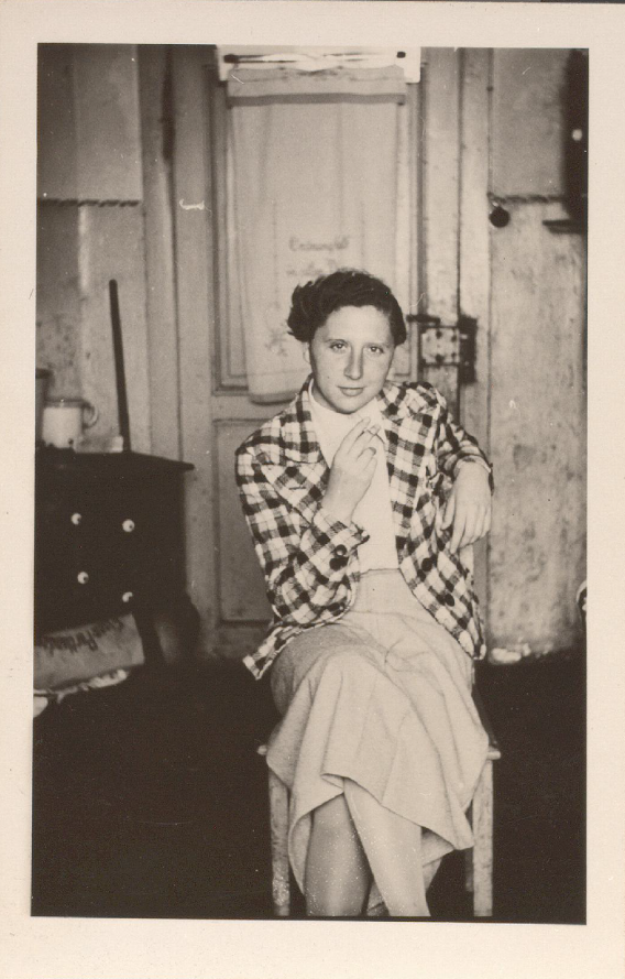 _Kohlmann sits on a chair in a private apartment in a skirt, blouse and checked blazer, a cigarette in their hand.