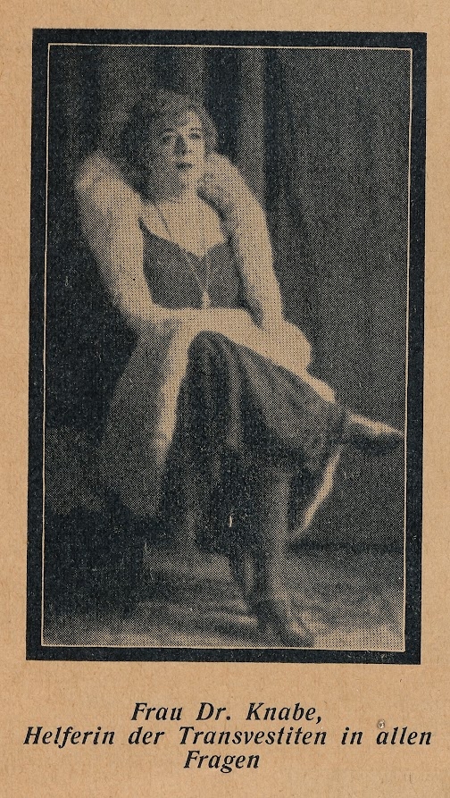 Knabe sitting on chair in dress, necklace, short hairstyle and coat with fur collar, legs crossed, caption: Frau Dr. Knabe, helper of the transvestites in all questions.