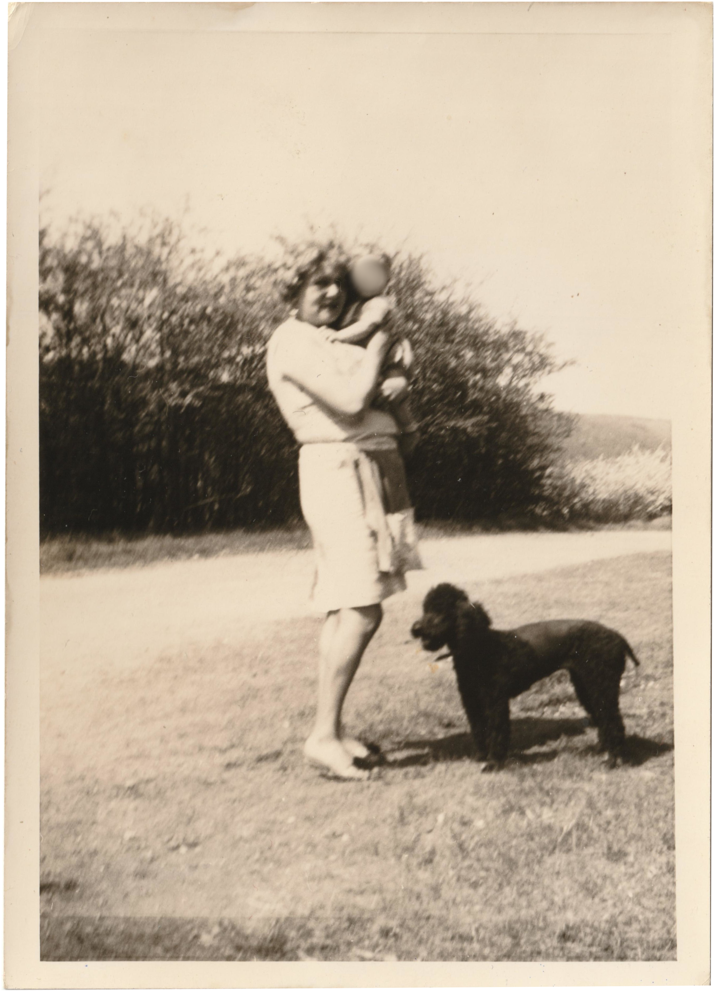 Private photo owned by Adeles niece Anni, Adele Haas in a skirt and blouse on a meadow, she is holding a small child in her arms, a black poodle stands in front of her.