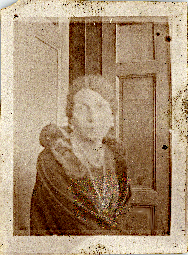 Charlotte Charlaque profile photo, she is sitting in a corner, probably in a private apartment, with doors on each side in the background. She wears round earrings, a long necklace, and a cloak with a fur collar.