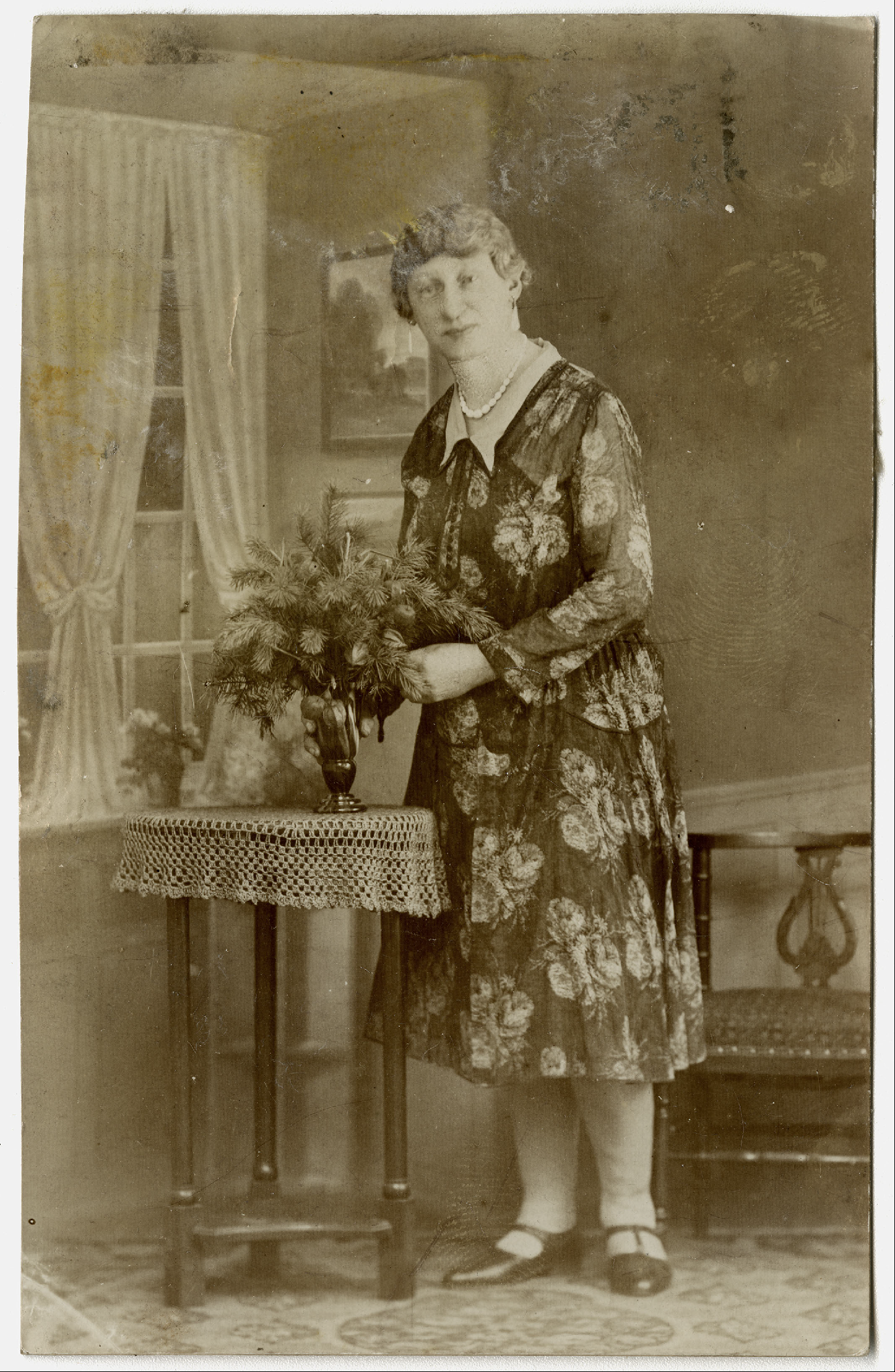 Emi Wolters stands on carpet in floral dress with light collar and pearl necklace. In front of her a small table with flower vase containing pine branches. Emi touches vase and branches. Taken in photo studio, windows and wall paneling are paintings.