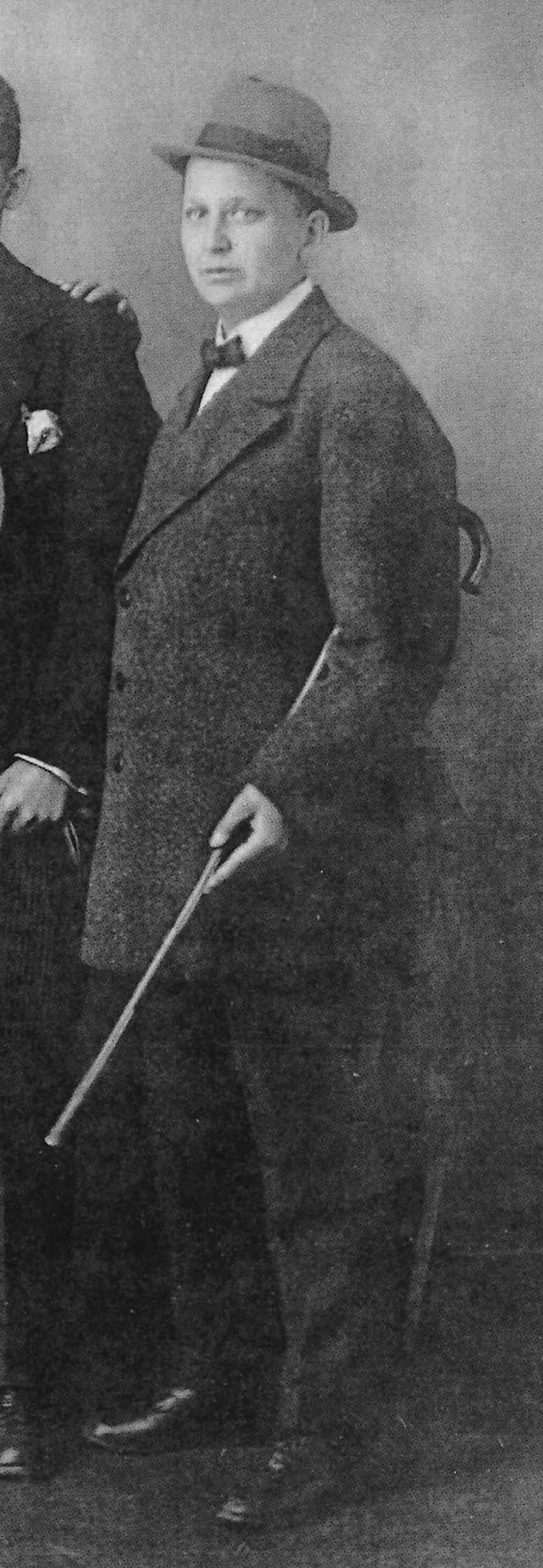 cut out part of an image, Ernst Kubbe stands on the right side of the photo in a shirt, tie, suit and hat and holds a cane under his arm.